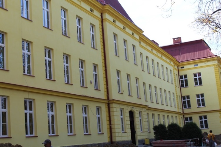 Trzykondygnacyjny budynek z żółtą elewacją i czerwonym dachem. Liczne prostokątne okna rozmieszczone równomiernie. Główne wejście z łukowym portalem. Przed budynkiem ławki i krzewy ozdobne.